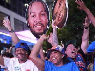 Crowd of cheering sports fans at night, wearing jerseys and caps, raising hands and holding large cutouts of a basketball player.