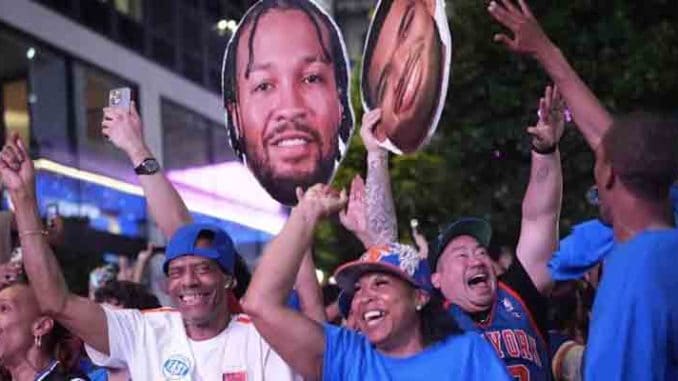Crowd of cheering sports fans at night, wearing jerseys and caps, raising hands and holding large cutouts of a basketball player.