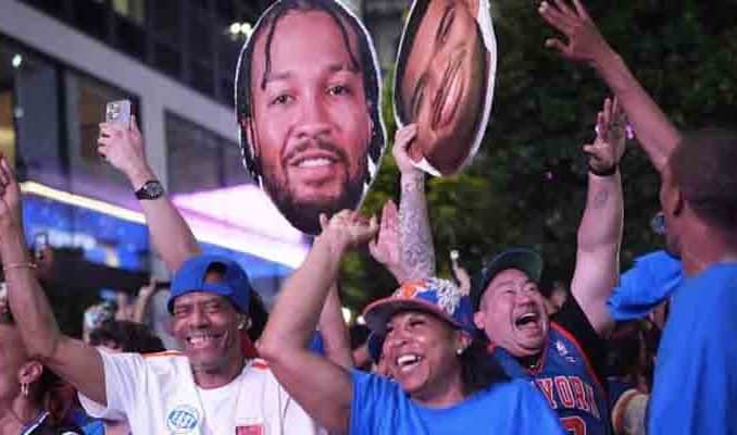 Crowd of cheering sports fans at night, wearing jerseys and caps, raising hands and holding large cutouts of a basketball player.